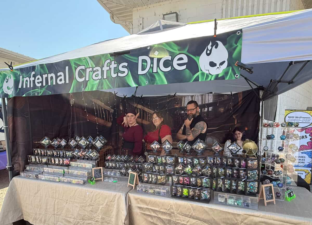 Infernal Crafts Dice booth at Magic Valley Comic Con 2025, featuring the dice smiths posing behind displays of handmade resin dice under a green banner.